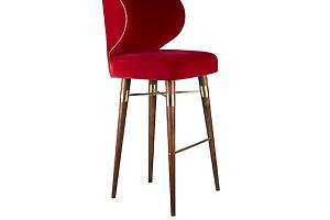 A high-resolution photograph of the Louis bar chair by Ottiu, showcasing its profile view. The chair features an upholstered body in a luxurious deep red velvet, bordered by a thin metallic piping that follows the curve of the crescent-shaped backrest. Its tall, tapered legs and support frame are crafted from rich, dark smoked walnut wood. Polished brass connectors and footrests bridge the wooden legs, adding a modern and elegant accent. The chair is positioned to clearly display the side profile of the seat, back, and the complex leg structure.