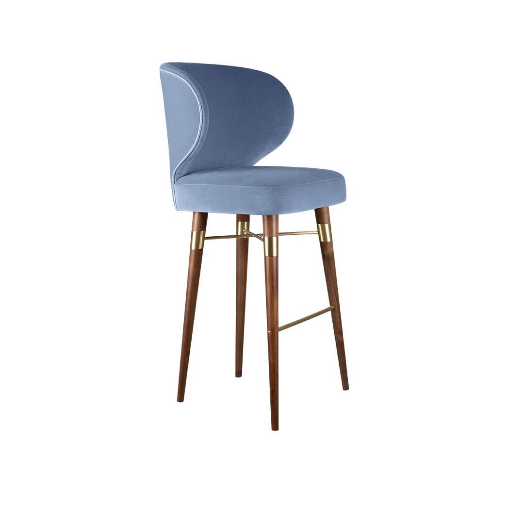 A high-resolution photograph of the Louis bar chair by Ottiu, showcasing its profile view, but upholstered in a soft, medium-blue velvet. The chair features the same crescent-shaped backrest with delicate metallic piping and sits upon dark smoked walnut legs. Polished brass details are present on the leg connectors and footrest, offering a sophisticated contrast to the blue upholstery. This image provides a clear look at the alternative color option, allowing for visualization of the piece in different interior schemes while maintaining the identical structure.