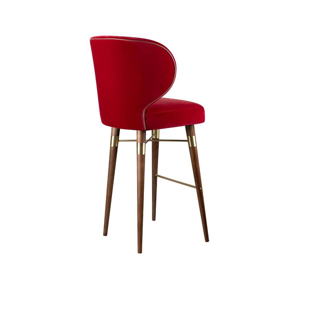 A high-resolution photograph of the Louis bar chair by Ottiu, captured from a rear three-quarter angle. This perspective highlights the elegant curve of the deep red velvet backrest and the tailored detail of the metallic piping that frames it. The dark smoked walnut legs are depicted in a dramatic angle, showing the depth of the leg structure. Polished brass components on the upper frame and lower footrest catch the light, providing a subtle sheen that contrasts with the rich wood and velvet textures.