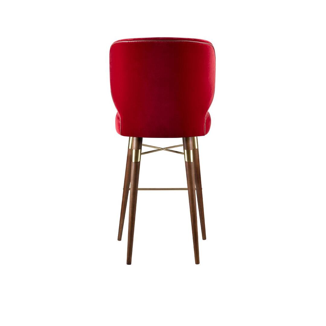 A high-resolution photograph of the Louis bar chair by Ottiu, presented in a straight-on rear view. This angle emphasizes the solid, curved shape of the deep red velvet backrest from the back, showing its form and texture. All four smoked walnut legs are visible, extending down with a slight taper. The complex arrangement of polished brass cross-bracing and the footrest connecting all legs is clearly displayed, providing a view of the chair’s structural support and detailing.