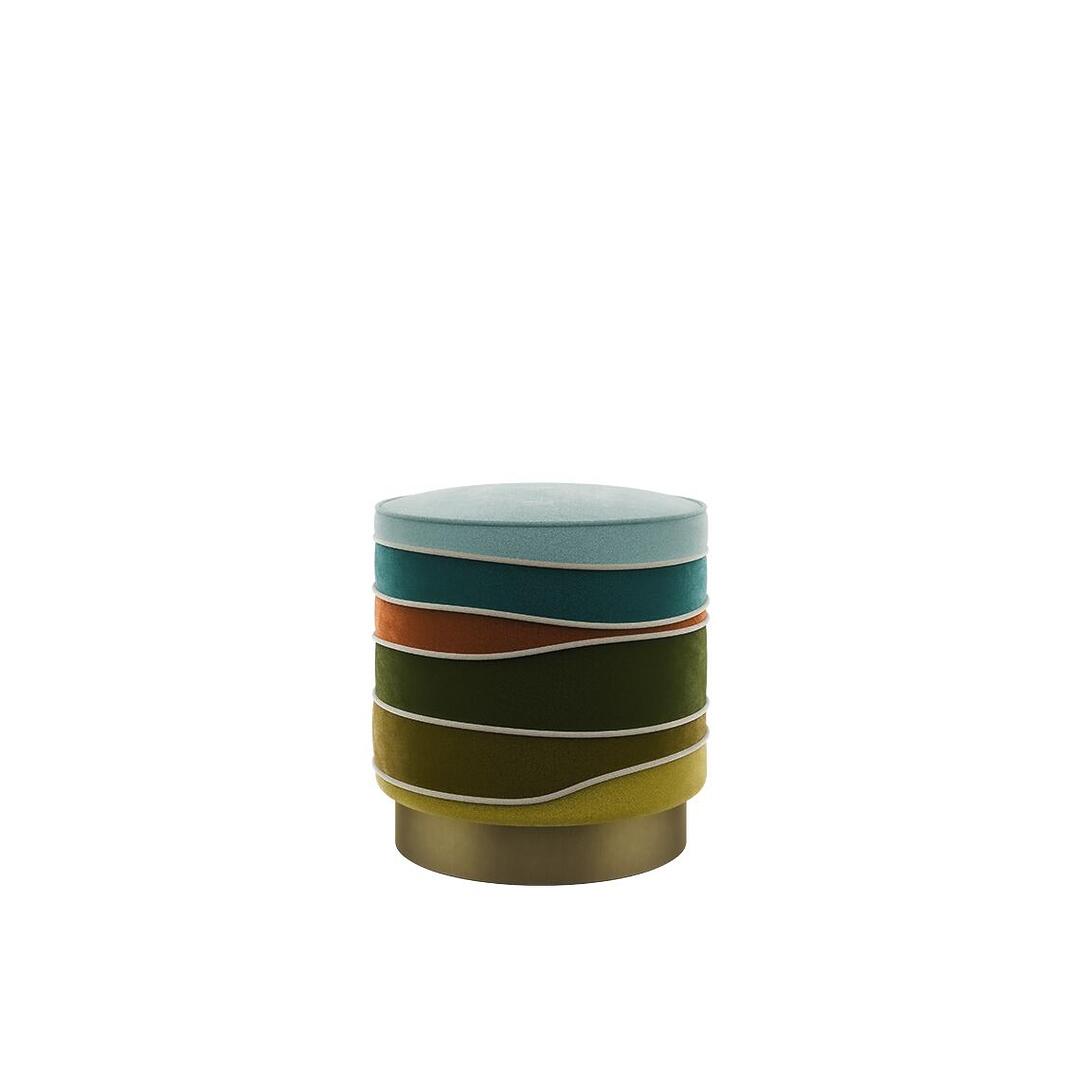 A front-facing studio view of the Charisse stool by Ottiu. The design showcases wave-like horizontal segments in a vibrant velvet palette of light blue, teal, orange, and various shades of green. Thin velvet piping accentuates the organic curves of each section, all resting on a cylindrical base made of brushed brass.