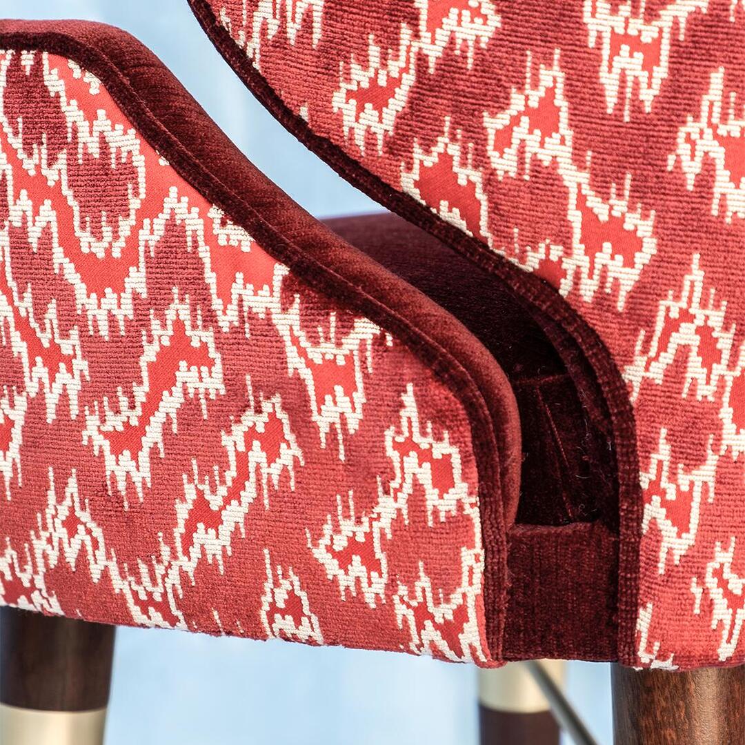 A close-up view of the Ottiu Louis I bar chair, detailing the patterned Schumacher Antique Strie Velvet in redwood and the solid dark red velvet piping. The top of the dark wood legs with brushed brass accents is visible.