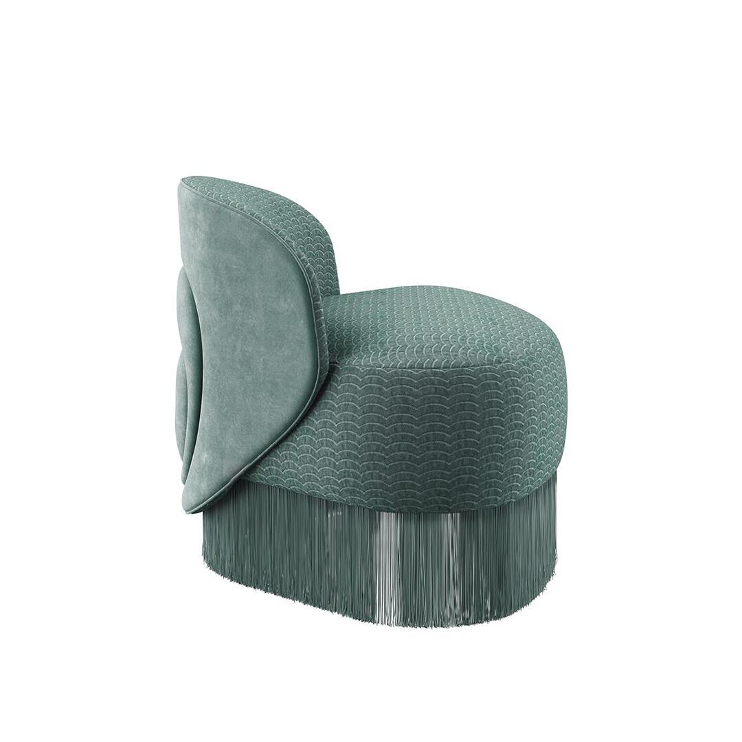 Doris armchair shown from the side. The piece highlights a lavish composition, exquisitely hand-upholstered in sumptuous turquoise velvet fabrics, elegantly draped with cascading fringes that exude grandeur. Upholstered in Dedar fabrics: Ripley and Plushy.