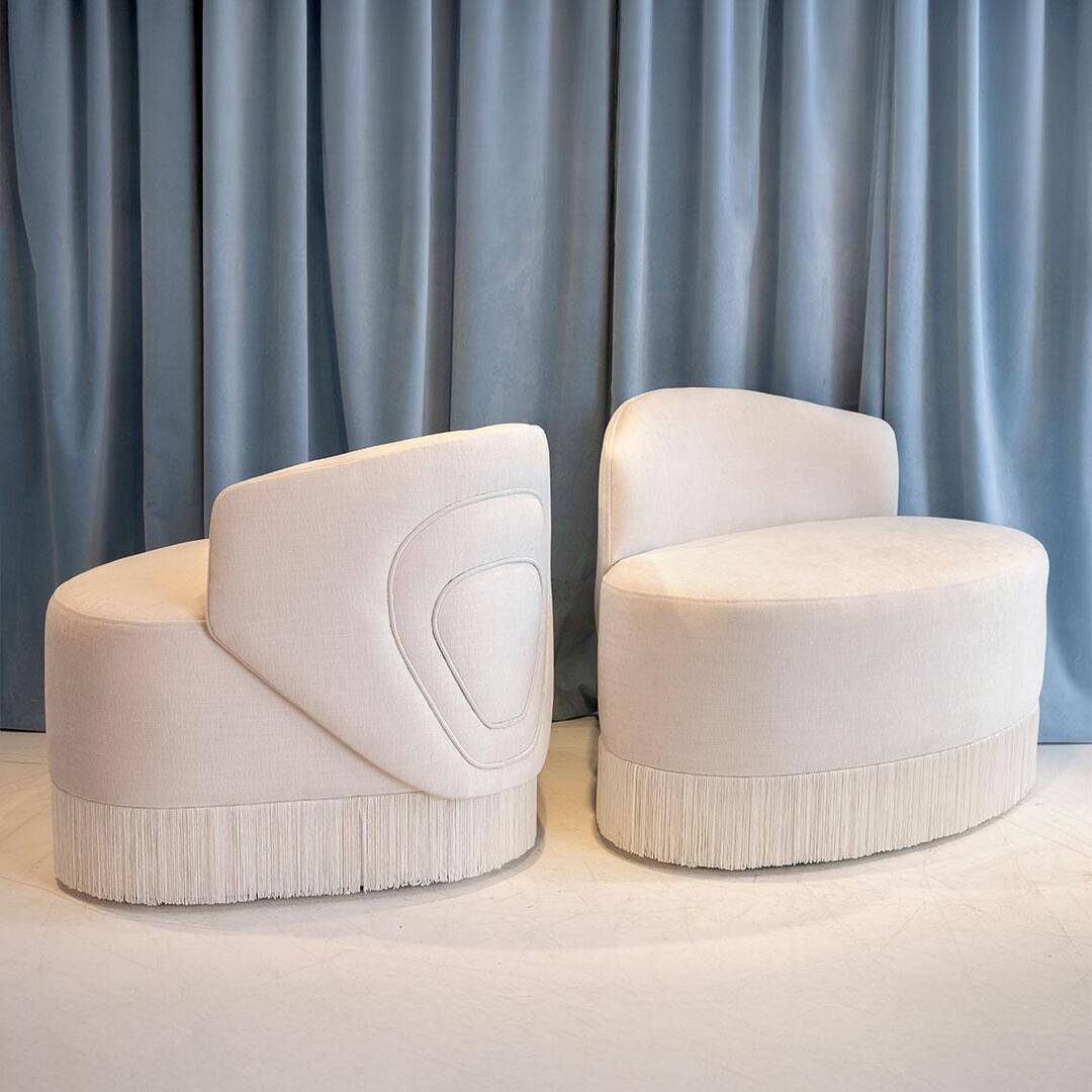 Two white Doris Armchairs upholstered in chenille with tricotine fringes, positioned at 45 degrees facing and backing each other.