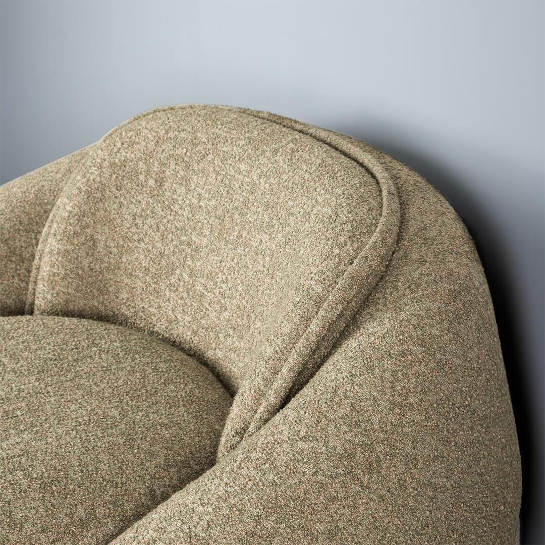 A close-up, high-angle shot of the Romy Armchair backrest and seat. The chair is upholstered in a textured, multi-tonal beige and olive green bouclé fabric. The image highlights the rounded, organic silhouette of the backrest and the soft, plush cushioning of the seat against a plain light gray wall.