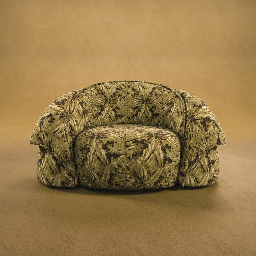 A front-facing studio shot of the Romy Armchair featuring a bold, camouflage-inspired pattern in shades of moss green, tan, and dark brown. The armchair has a low-profile, wide-set design with oversized, rounded armrests and a deep seat. It is centered against a textured warm ochre background.