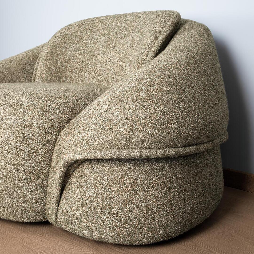 A detailed side view of the Romy Armchair in textured bouclé fabric, showcasing its unique sculptural construction. The design features a continuous, wrap-around upholstered panel that creates the armrest and base, separated by a deep horizontal seam. The chair is positioned on a light wood floor against a neutral wall.