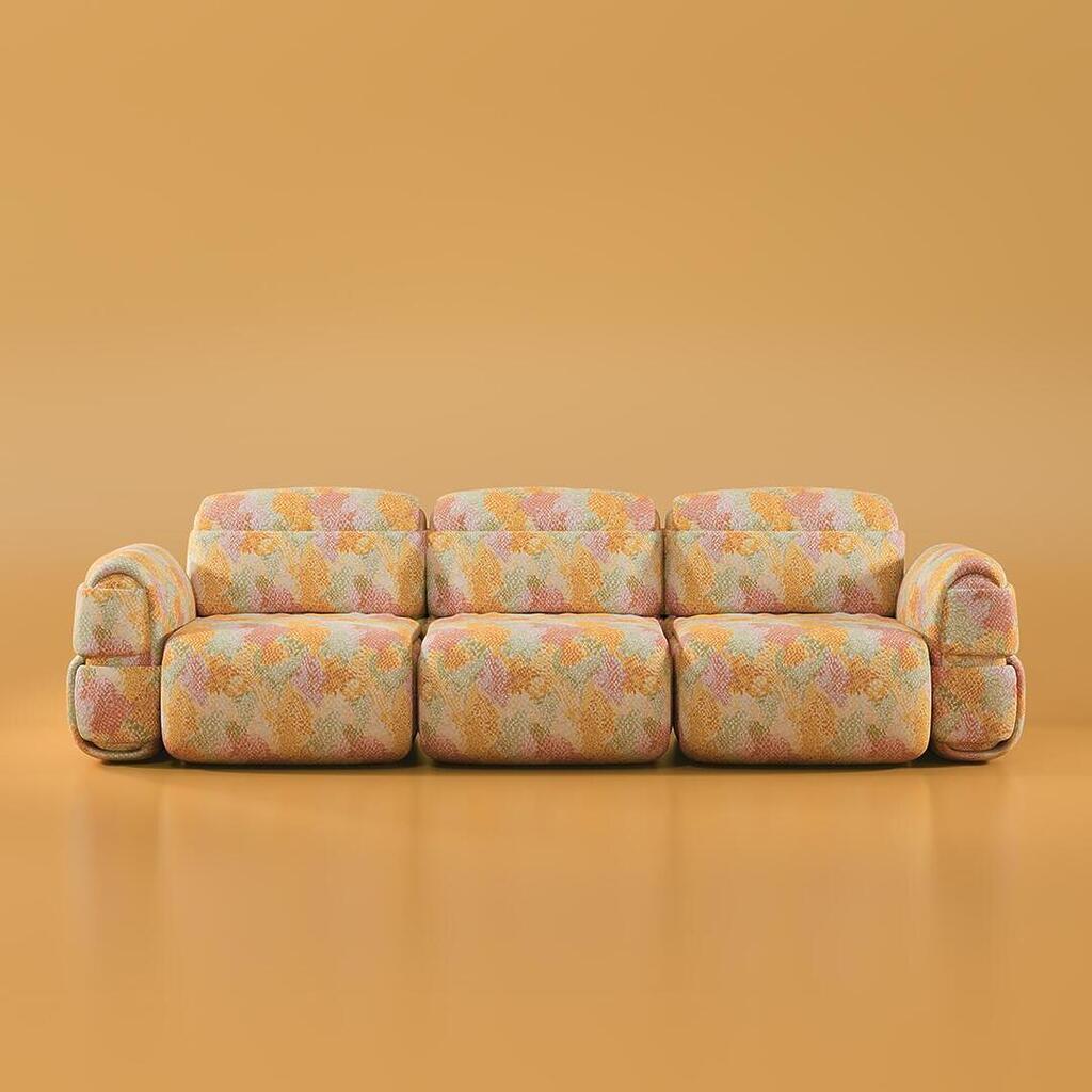 Front-facing view of the Elsa Modular 3-seater sofa, showcasing its voluminous silhouette and soft horizontal padded rolls. The sofa is wrapped in a vibrant abstract patterned fabric against a reflective caramel-colored studio backdrop.