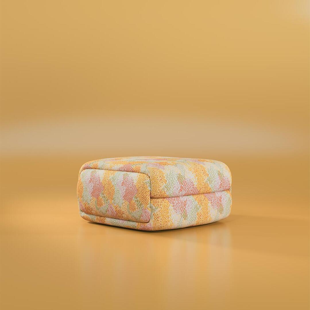 A close-up perspective of the Elsa Modular Ottoman, designed with soft edges and a padded texture. It is covered in a multicolored textured fabric with a painterly pattern, resting on a polished ochre-toned floor.