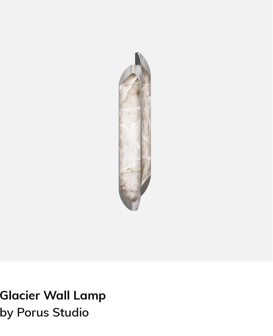 The Glacier Wall Lamp, a sleek and modern vertical sconce. It features a slim, capsule-shaped polished metal frame that houses two layered panels of translucent white stone with natural grey veining. When lit, the stone panels create a soft, diffused glow, emphasizing their organic texture and the lamp's minimalist architectural design.