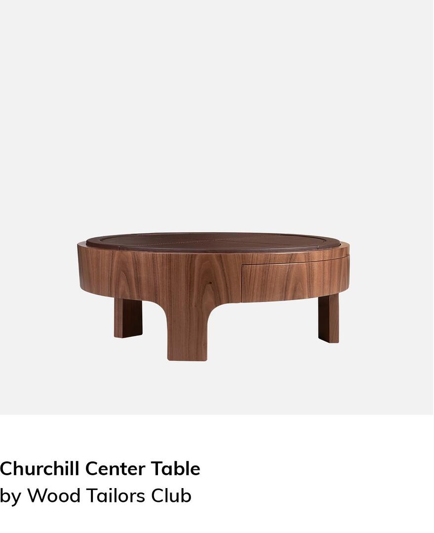 The Churchill Center Table, a low circular coffee table crafted from polished walnut. It features a thick, rounded frame with an integrated drawer and a dark brown leather-inlaid top with cross-stitching detail. The piece is supported by three sturdy, curved wooden legs that seamlessly blend into the table's base.