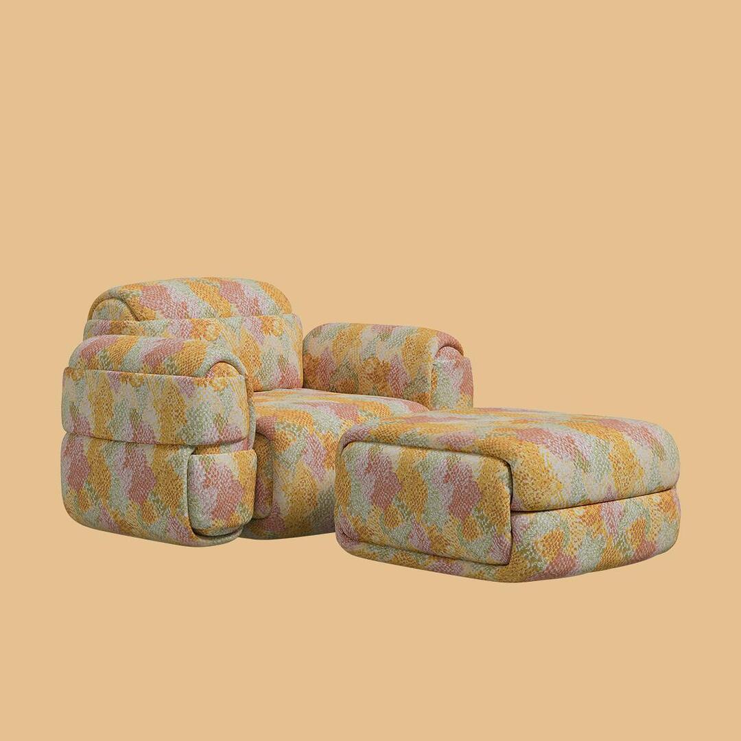 The Elsa Modular Armchair I paired with the Elsa Ottoman. The setup includes a voluminous armchair with thick, rounded armrests and a matching rectangular pouf placed directly in front of it. Both pieces are uniformly upholstered in a textured, brightly colored fabric featuring a mosaic pattern of yellow, coral, green, and peach.