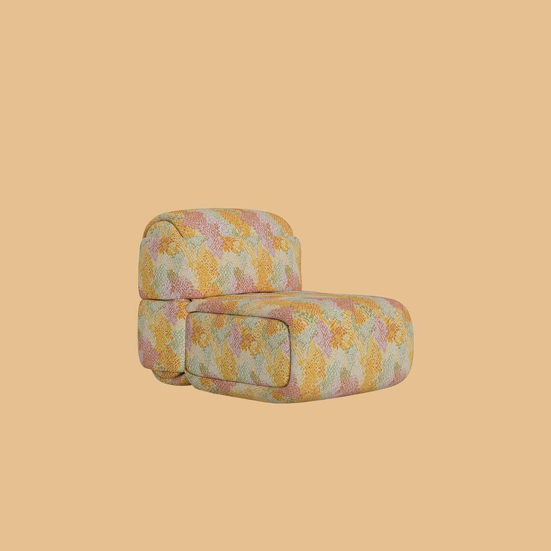 The Elsa Modular Armless Chair, a sculptural lounge seat featuring a thick, rounded cushion base and a matching curved backrest. It is fully upholstered in a textured fabric with a mosaic-like geometric pattern in warm shades of yellow, coral, peach, and sage green.