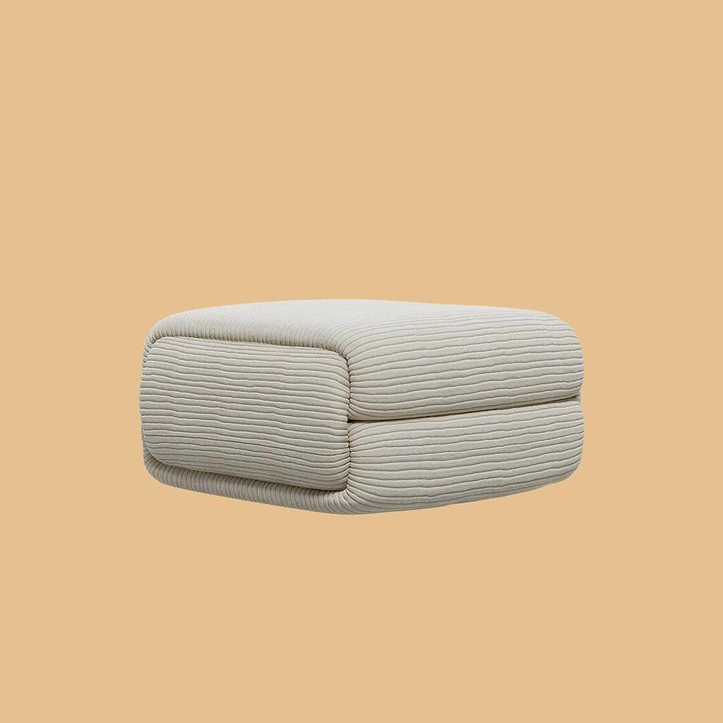 The Elsa Ottoman, viewed from an angle. The square, two-tiered pouf showcases a plush, sculptural silhouette and is entirely upholstered in an off-white, horizontally ribbed fabric.