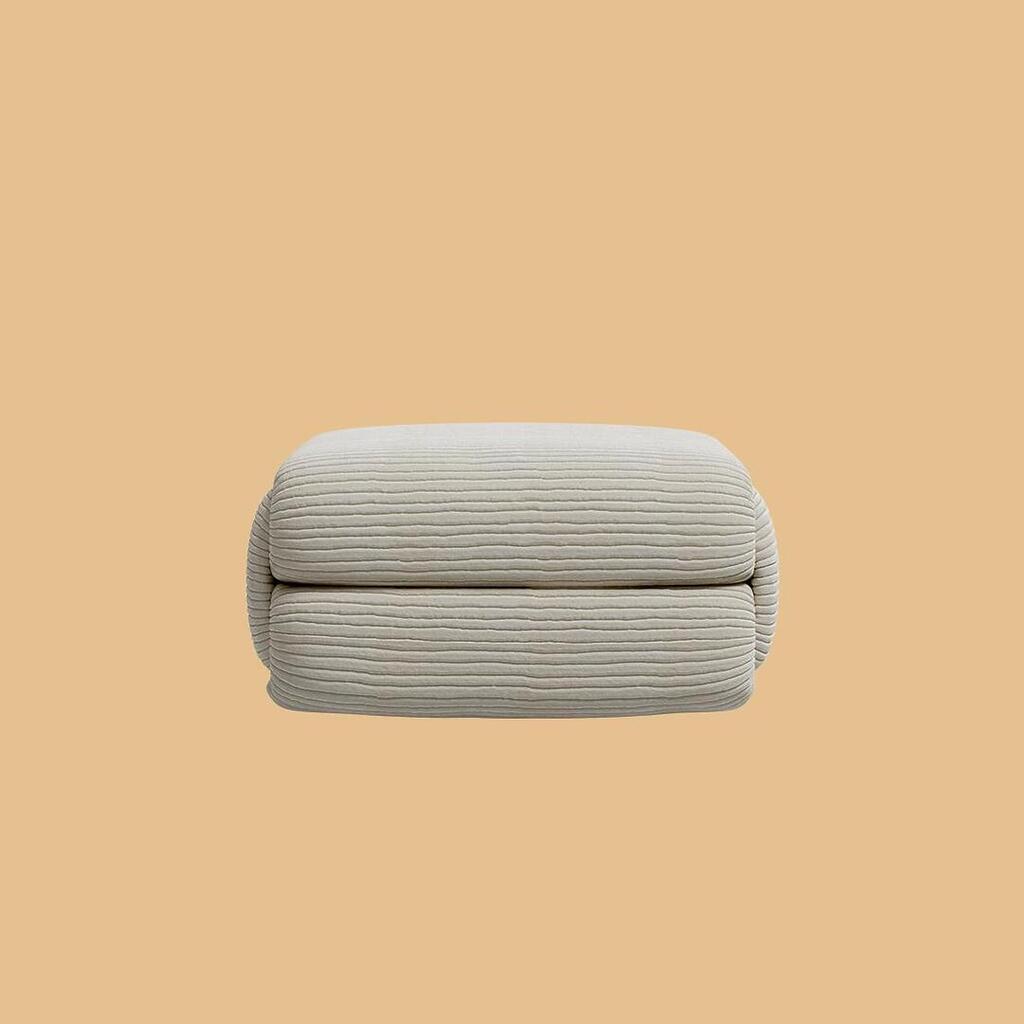 The Elsa Ottoman, viewed straight on. This profile highlights the plump, two-tiered segmented design of the square pouf, which is uniformly covered in a textured, off-white horizontally ribbed fabric.