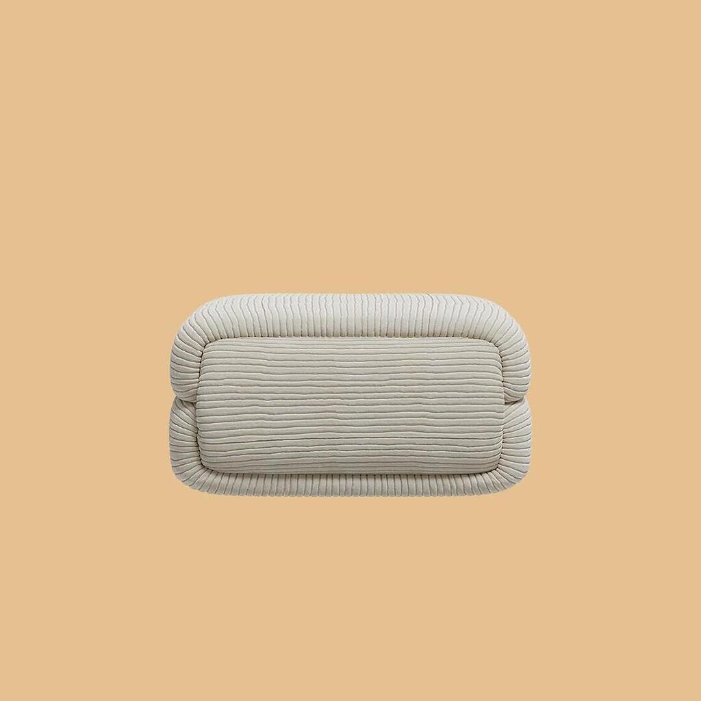 The Elsa Ottoman, viewed straight on. This profile highlights the plump, two-tiered segmented design of the square pouf, which is uniformly covered in a textured, off-white horizontally ribbed fabric.
