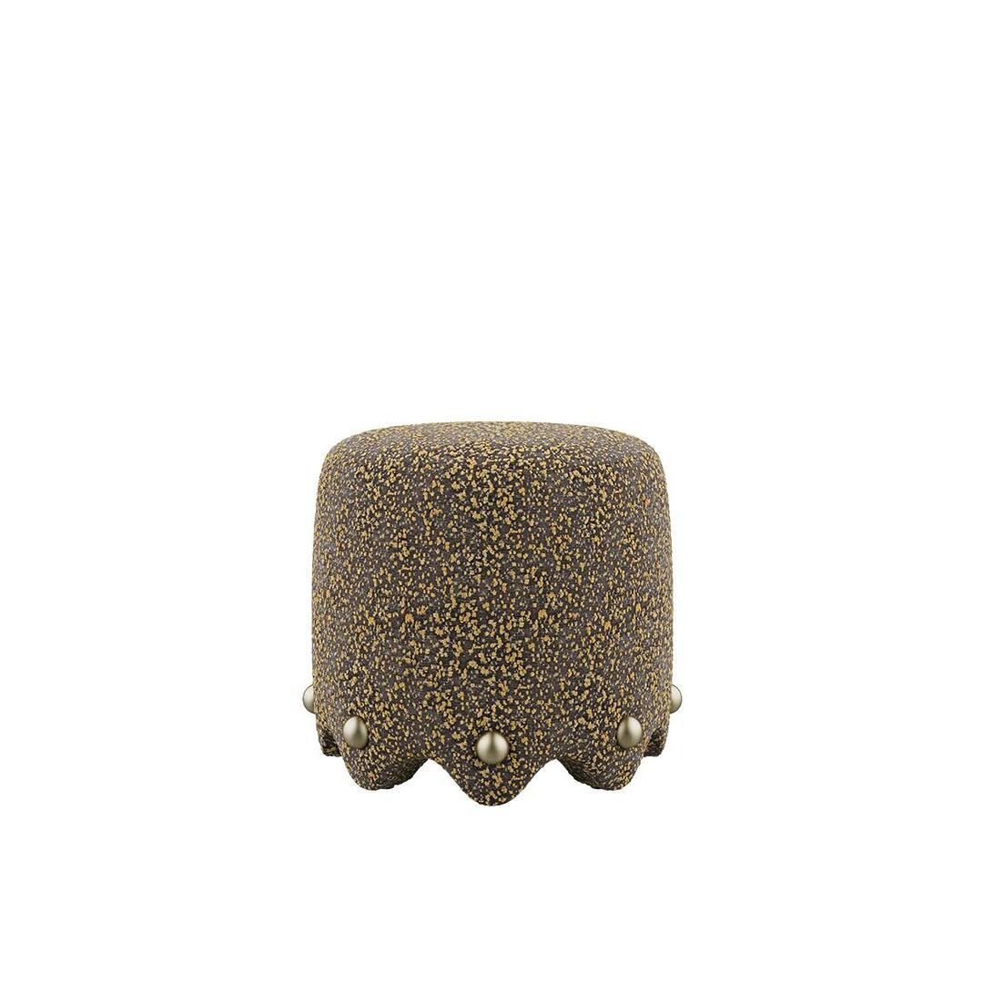 A variation of the Bette Stool by Ottiu, showcasing the assertive sculptural form in a textured, speckled ochre and charcoal Élitis Mochi fabric. The scalloped bottom edge is accented with golden brass spheres, creating a bold contrast between the soft textile and the metallic hardware.