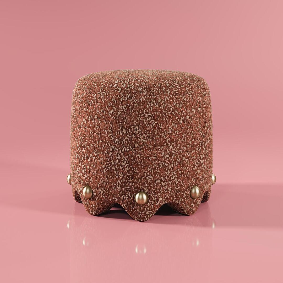 A lifestyle shot of the Bette Stool by Ottiu set against a soft pink background. The stool features a heavy, rounded silhouette upholstered in rust-toned Élitis Mochi fabric with a rich, nubby texture. At the base, a distinctive scalloped edge is decorated with polished brass spheres that reflect the ambient light on a glossy pink floor.