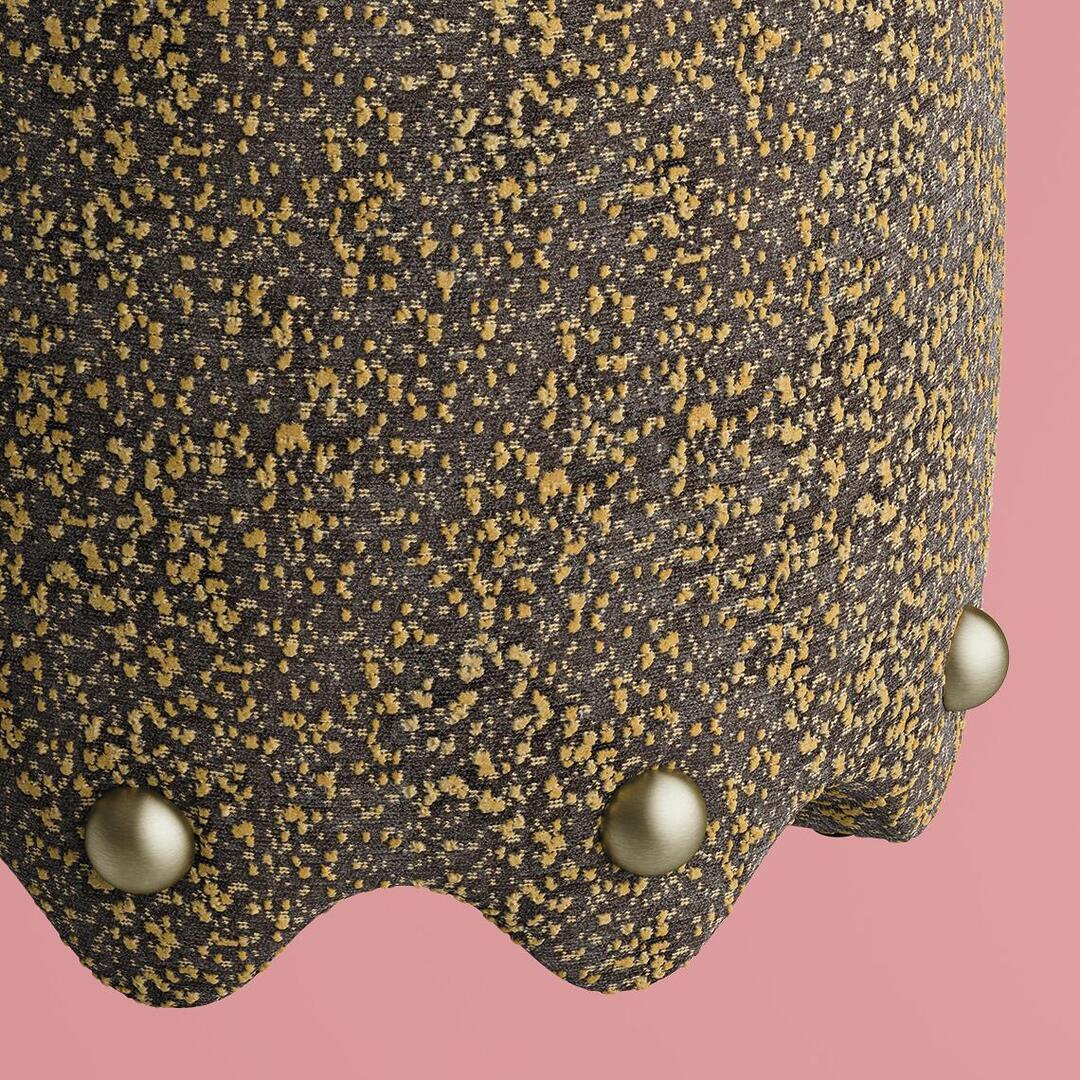 A detailed close-up of the Bette Stool by Ottiu, showcasing the intricate weave of the Élitis Mochi fabric in a charcoal and ochre colorway. The focus highlights the tactile, raised dots of the textile and the smooth, matte finish of the signature brass spheres embedded in the sculptural scalloped hemline against a pastel pink backdrop.