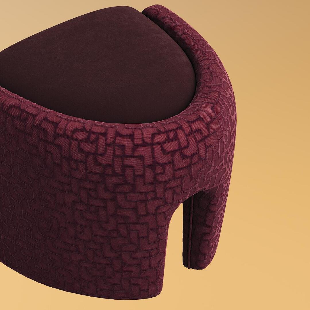 A high-angle, close-up shot of the Grace Stool by Ottiu in a rich burgundy colorway, set against a soft beige background. The image highlights the contrast between the plush, smooth matte-velvet seat cushion and the surrounding sculptural frame wrapped in a deeply textured, patterned burgundy velvet with subtle geometric engravings. A portion of the stool's elegantly tapered, arched leg is visible.