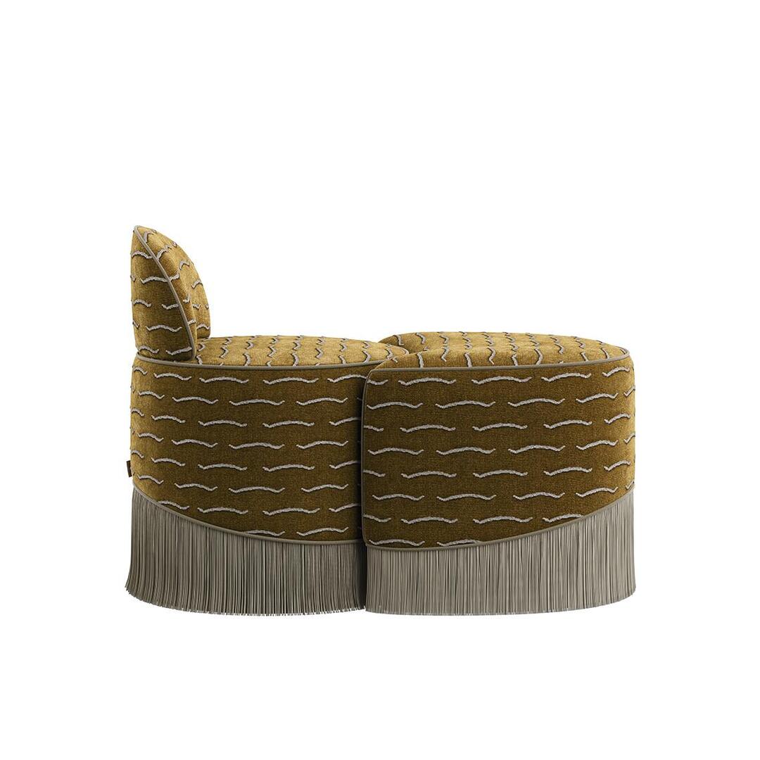 A direct side profile view of the Lila Stool by Ottiu. The image displays how the low backrest section with a rounded silhouette perfectly links with the crescent-shaped footrest section. The continuous horizontal wave pattern of the gold velvet fabric and the straight hang of the long cream fringe base are clearly visible.