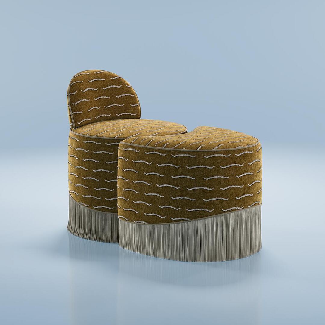 A studio shot of the Lila Stool by Ottiu, showcasing a modular, two-piece upholstered seating arrangement against a light blue background. The stools are wrapped in a mustard-yellow or olive-gold jacquard velvet fabric with a wavy, horizontal white pattern. One section features a low, curved backrest, while the matching ottoman section fits snugly against its curved front. Both pieces feature a long, cascading greyish-beige fringe trim at the base that touches the reflective blue floor.