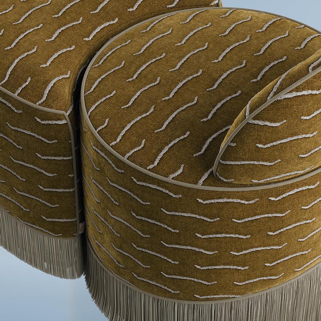 A close-up, high-angle detailed view of the Lila Stool by Ottiu. The shot highlights the rich texture of the mustard-gold jacquard velvet upholstery and the intricate, white wavy lines patterned across the surface. Greyish-beige piping neatly defines the edges of the modular seating sections and the rounded backrest. The dense, long fringe at the base is visible, creating a soft, textured border along the bottom of the curved furniture pieces.