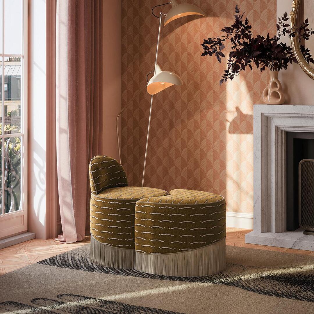 An elegant interior scene featuring the Lila Stool by Ottiu, a modular two-piece vanity stool and ottoman upholstered in a rich mustard-yellow jacquard velvet with a wavy white pattern and a dramatic, long fringe base. The sculptural stool is placed on a patterned rug in a sunlit room alongside a marble fireplace, a retro floor lamp, and warm-toned geometric wallpaper.