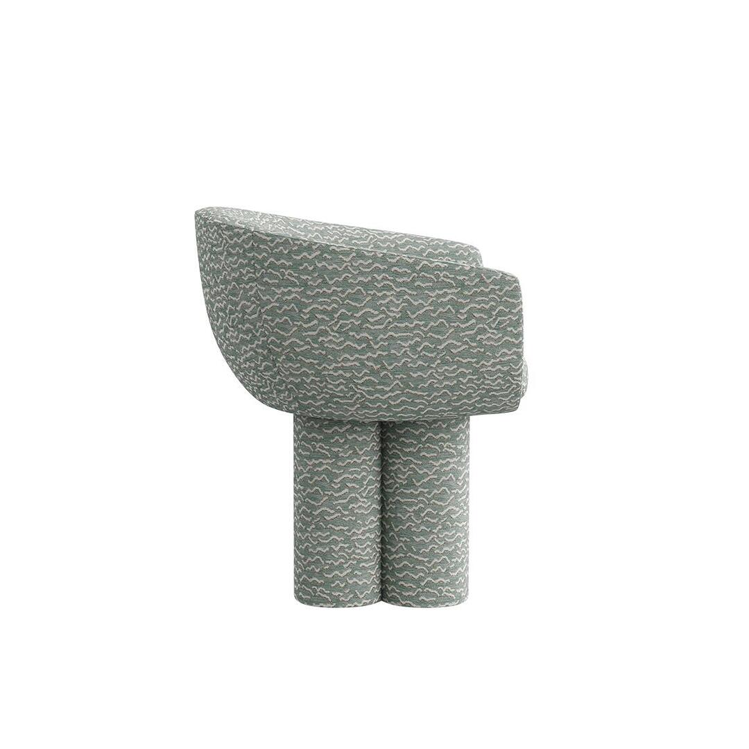 A side view of the Mae Dining Chair, with a direct look at the upholstered seat and the embracing backrest. The chair stands firmly on its singular leg column, all covered in the patterned "Pierre Frey - Le chant des dunes - Aqua" fabric,