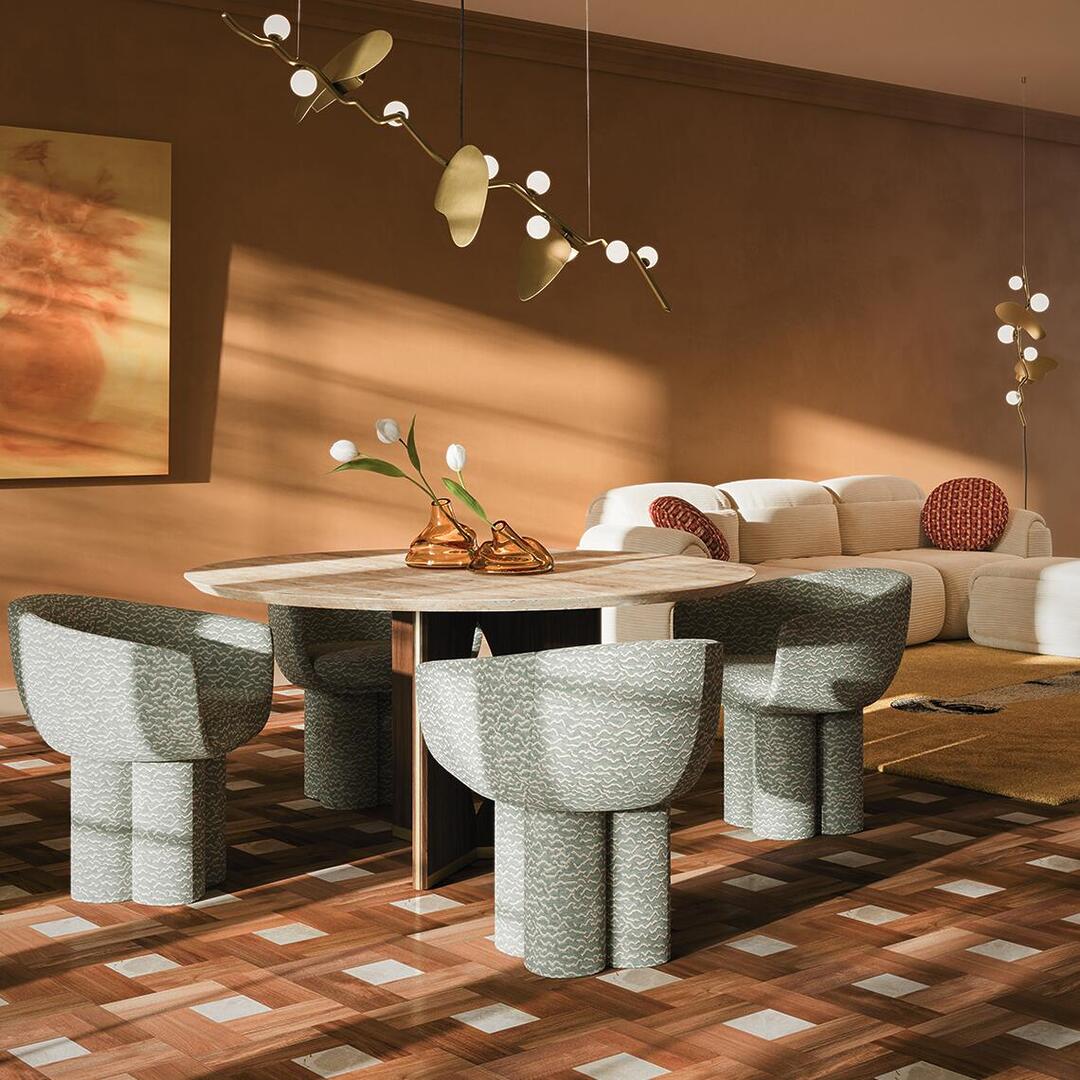A luxurious, warm-toned dining and living space featuring four Mae Dining Chairs by Ottiu upholstered in a textured aqua and cream pattern, surrounding the Taylor Dining Table by Wood Tailors Club. In the background, the Elsa Modular Sofa by Ottiu in a cream fabric is accented by reddish-patterned pillows. The scene is illuminated by the golden Almond Suspension Lamp and a matching Almond Pendant Lamp by Creativemary, which cast soft shadows across the terracotta-colored walls. The room is characterized by an earthy color palette of ochre, burnt orange, and rich wood tones from the patterned parquet floor, creating a sophisticated and inviting atmosphere.