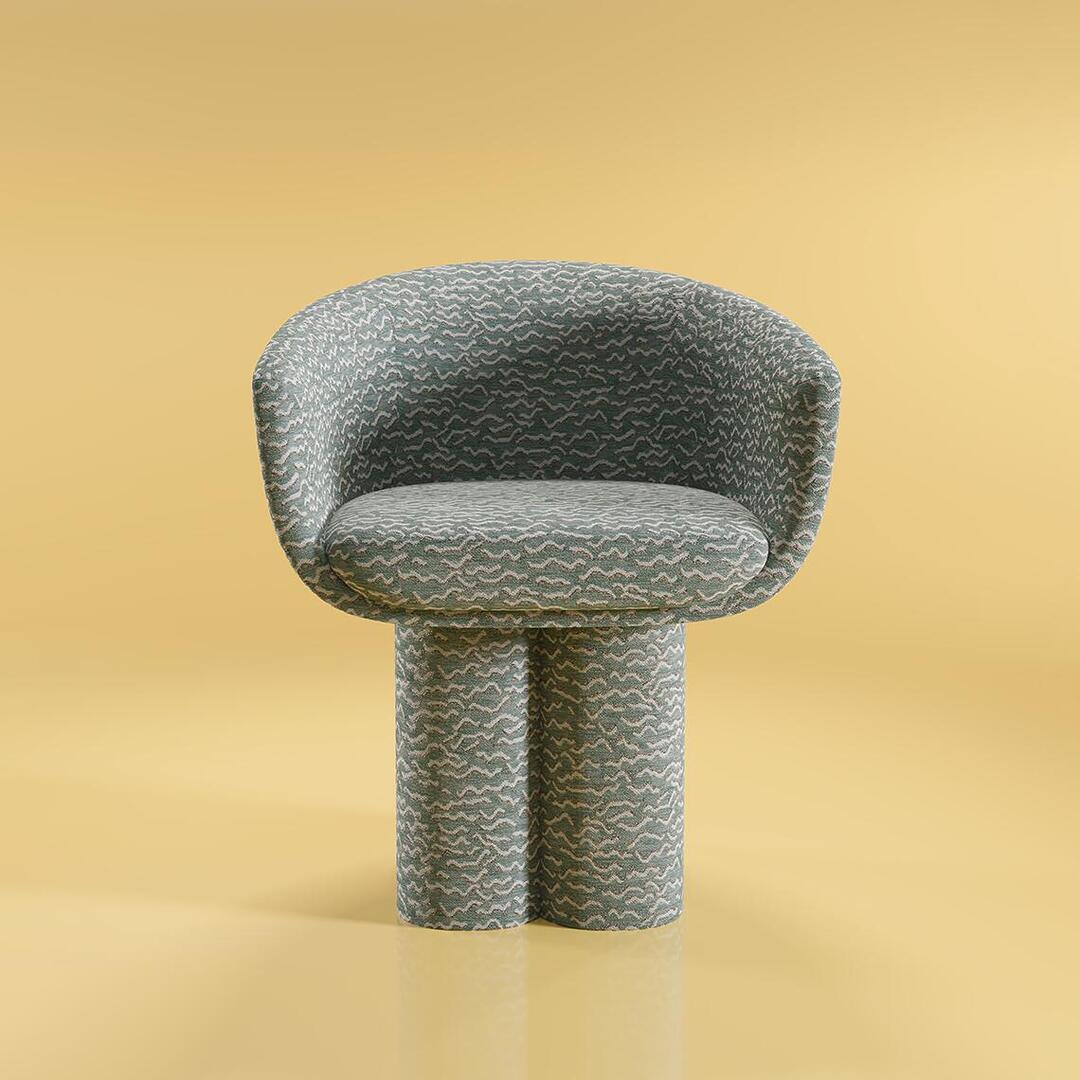 An interior studio photograph of the Mae Dining Chair, fully upholstered in "Pierre Frey - Le chant des dunes - Aqua" fabric, placed against a warm, muted yellow backdrop. The front-on perspective highlights the chair's rounded, sculptural form and the intricate fabric pattern.