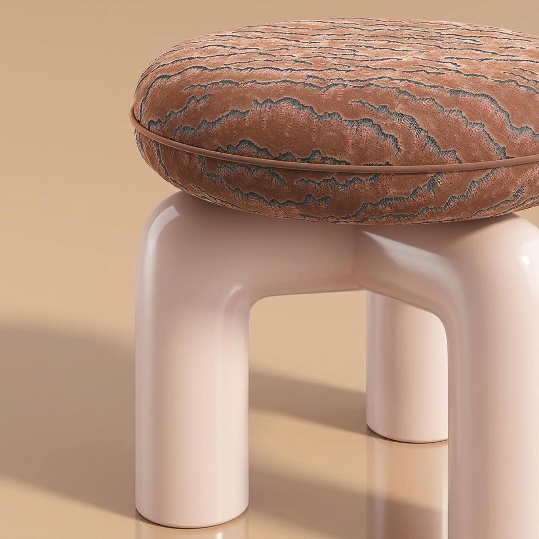 A detailed, close-up shot of the Tamara Stool by Ottiu, showcasing the rich textures and finishes. The round, plush seat is upholstered in a textured, multi-dimensional velvet with a brownish-pink and dark grey embroidered pattern resembling organic waves or bark. A matching velvet piping trims the center edge of the cushion. The seat rests on a glossy, pale pink lacquered wood base featuring thick, smooth, tubular arched legs that meet in the center. The stool is set against a warm, neutral beige background, with its glossy surface casting soft reflections on the floor.