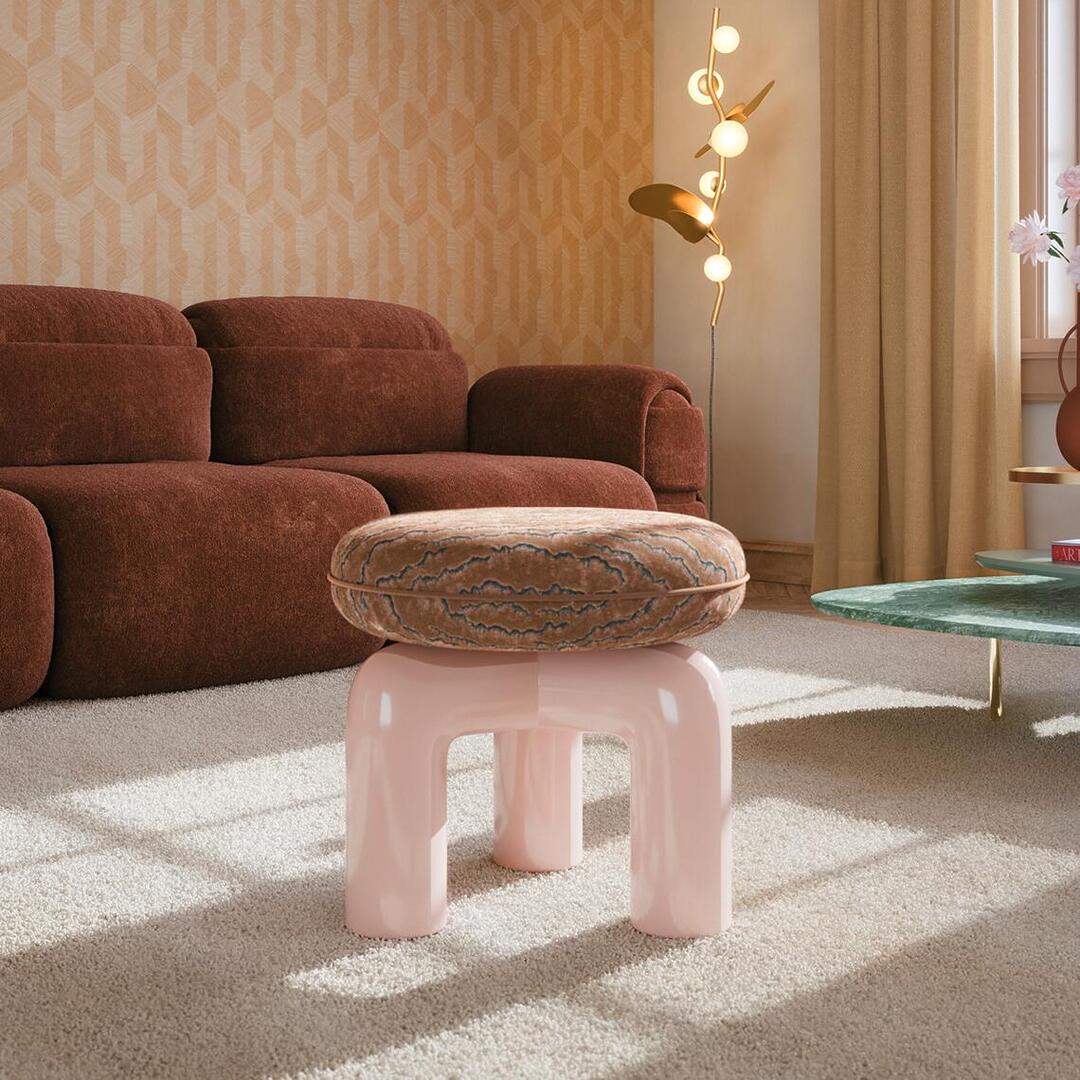 A contemporary, sculptural Tamara Stool by Ottiu stands in the foreground of a warmly lit, modern living room. The stool features a glossy, pastel pink lacquered wood base composed of thick, rhythmic tubular arches forming its legs. Atop the base sits a plush, rounded cushion upholstered in a rich, textured velvet with an intricate brown and blue embroidered pattern, finished with precise piping along the edge. In the background, a low-slung, textured rust-brown sectional sofa rests against a wall with geometric, light orange patterned wallpaper. Soft sunlight streams across a plush cream rug, illuminating the stool and casting long shadows, while a gold, organic-shaped floor lamp with glowing spherical bulbs stands in the corner near elegant beige curtains.