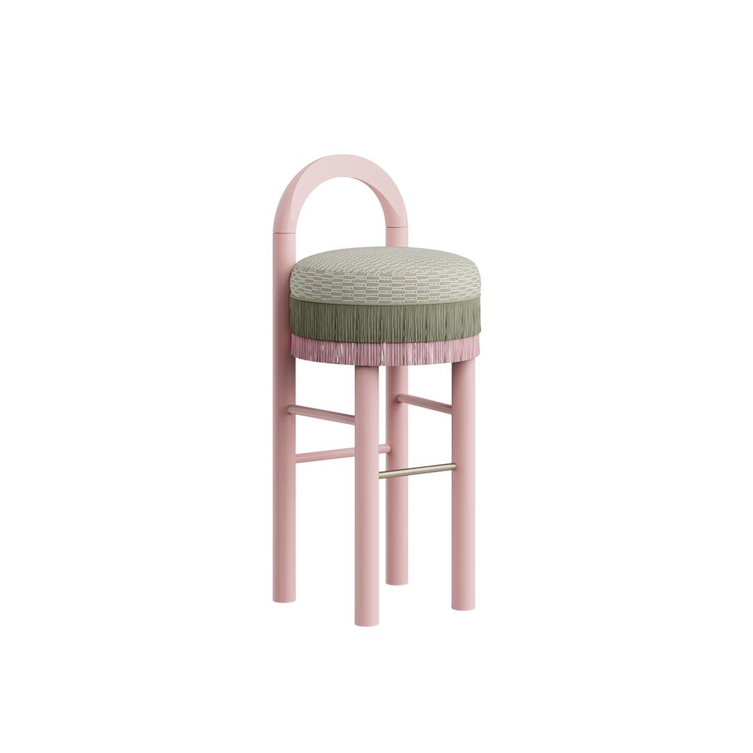 Three-quarter view of the Ann Bar Chair with soft pink lacquered wood legs, a geometric patterned upholstered seat cushion, green and pink two-tone fringes, and a stainless steel footrest.