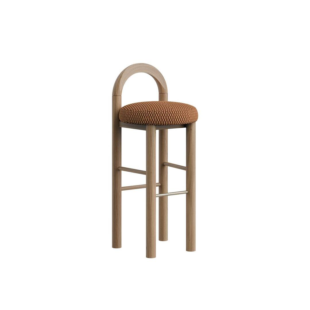 Three-quarter view of the Ann Bar Chair in natural wood with a checkered decorative weave upholstered seat cushion, arched backrest, and stainless steel footrest, shown without fringes.