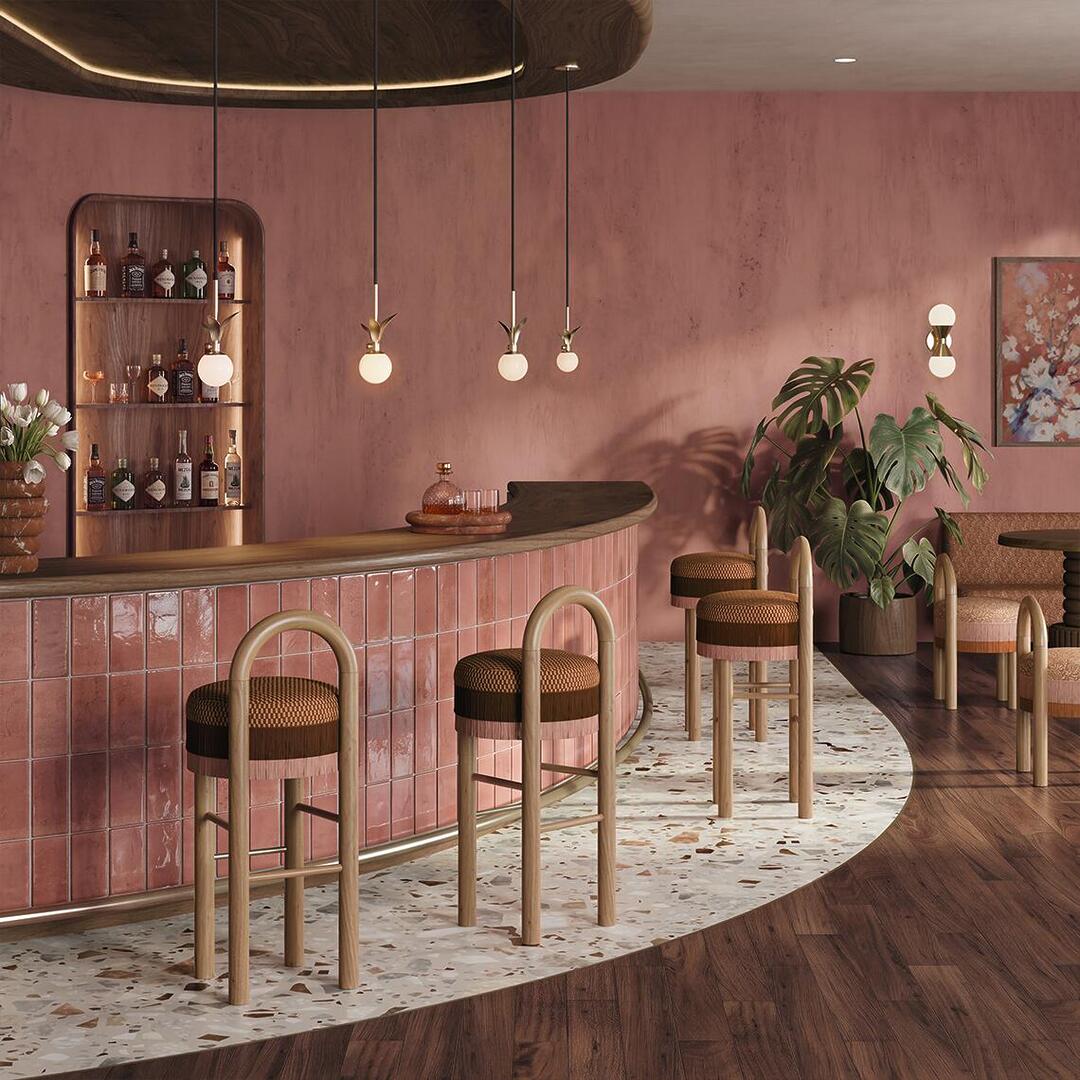 Stylish bar interior featuring Ann Bar Chairs with natural wooden legs along a curved counter with glossy pink tiles, terrazzo flooring, warm textured walls, pendant lighting, and tropical plants.