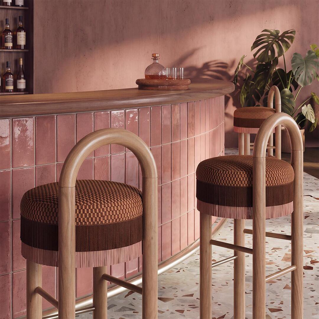 Close-up view of Ann Bar Chairs with natural wooden frames and fringe details placed along a curved pink tiled bar counter in a chic lounge setting.