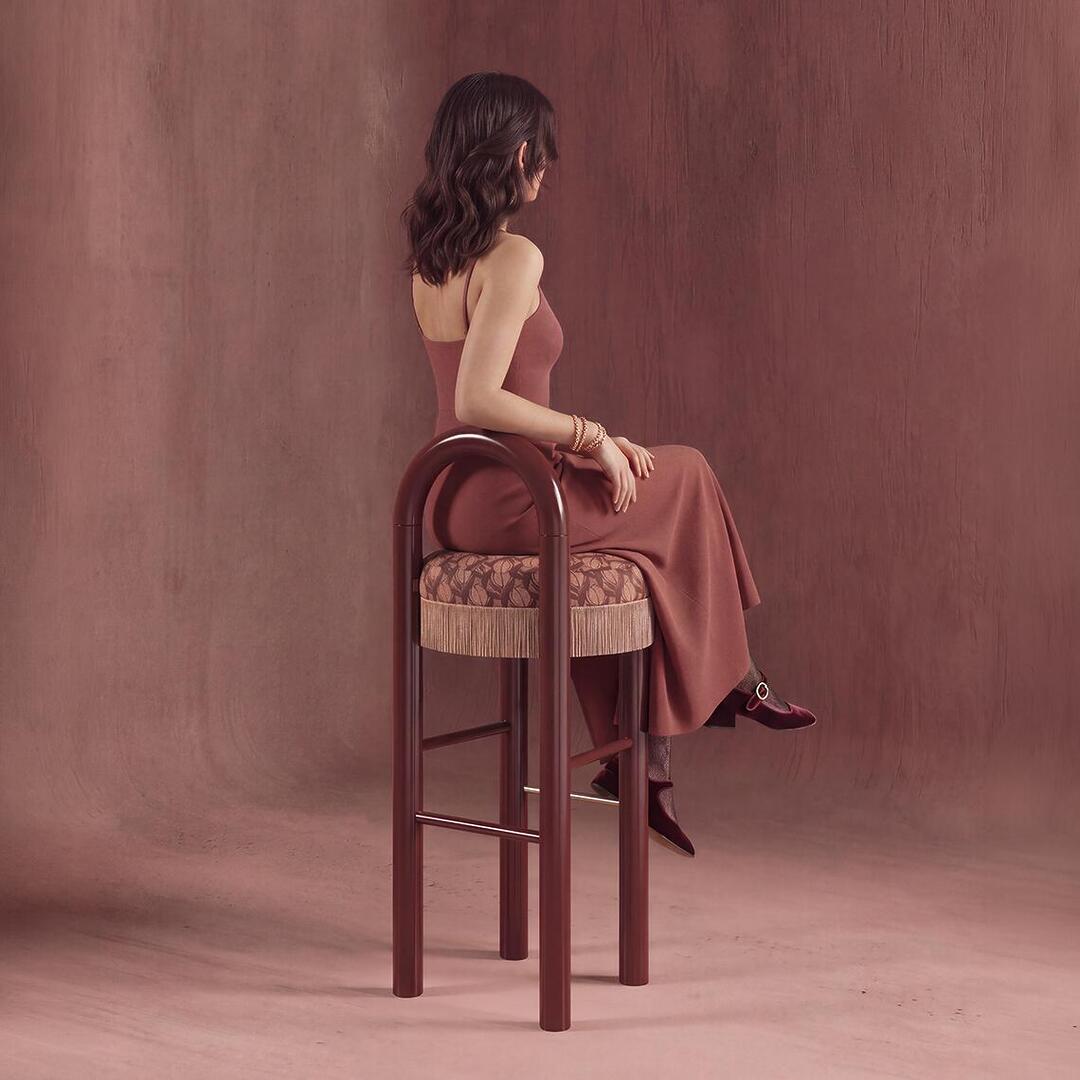 A woman in a rose dress seated sideways on a dark lacquered wood Ann Bar Chair against a matching warm textured backdrop.