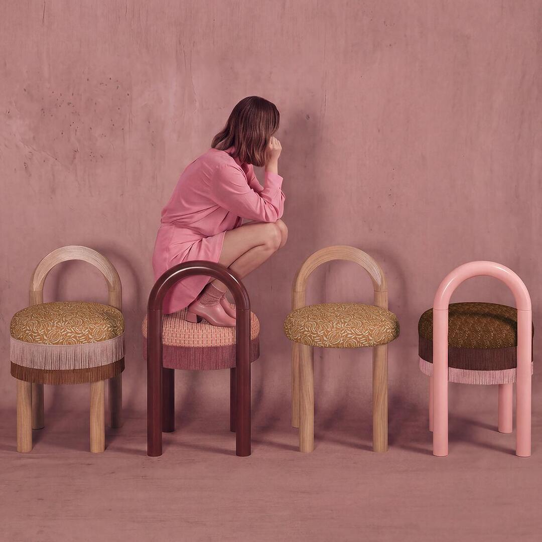 A stylized photo shows four Ann dining chairs in a row against a textured mauve background. A woman in a pink dress crouches on the second chair, looking back at the first. The stools are similar in form but feature different color combinations of legs, upholstery, and fringe.