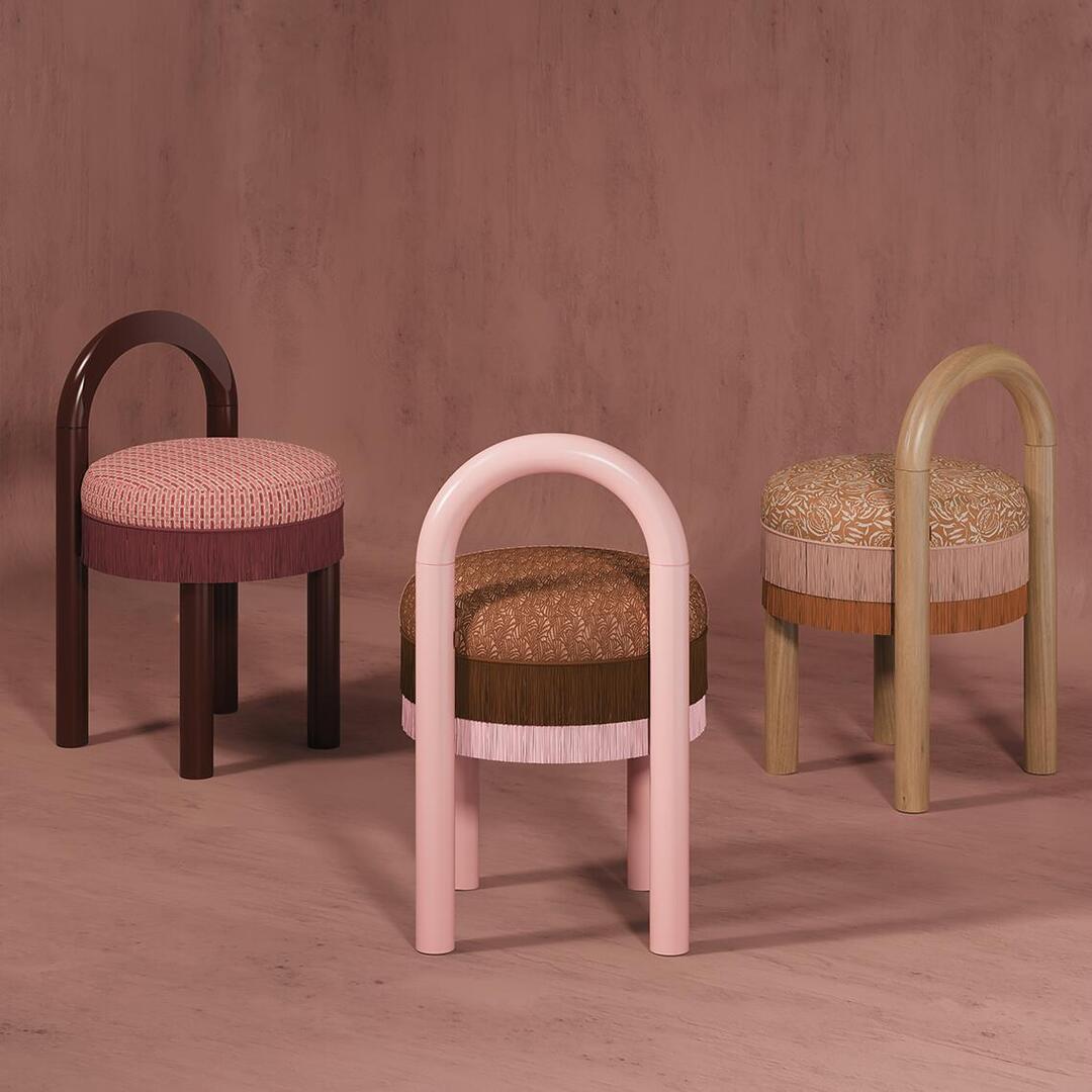 Three Ann dining chairs arranged on a dusty rose floor in a triangular composition. The stools showcase three distinct colorways: a brown-legged stool with a pink seat, a pink-legged stool with a brown seat, and a light-wood-legged stool with an orange seat, all with tiered fringe details.