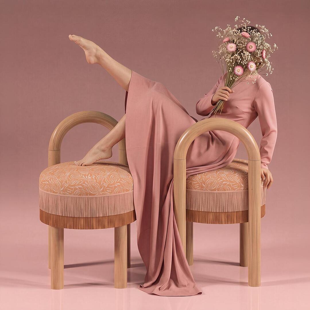 A high-fashion conceptual photo shows a model in a floor-length pink gown draped over two rounded wooden stools with fringed, patterned seats. Her face is obscured by a bouquet of dried pink and white flowers, and her bare foot is raised in an elegant, leg-up pose against a matching dusty rose background.