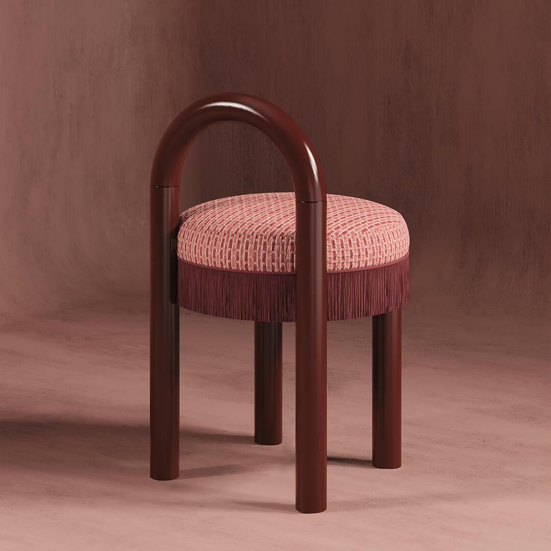 A studio product shot of Ann dining chair featuring an arched backrest and legs made of a deep maroon, high-gloss material. The round seat is upholstered in a textured pink and reddish weave and finished with a long, maroon-colored fringe. The backdrop is a textured mauve plaster wall.
